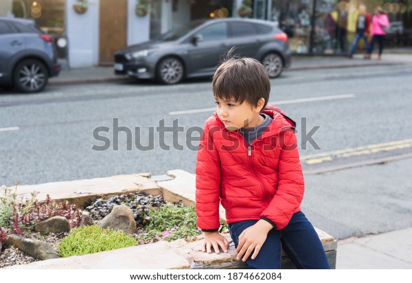 Lost Kid sitting alone next to a busy street in a\
center of city, Young boy with worrying face looking looking out\
deep in throught with blurry car on the road background,Lost\
children concept