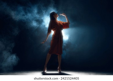 Lost in melody, bathed in light. Elegant and artistic woman in dress singing on stage against black background in dramatic smoke. Concept of hobby, festival, concert, disco, entertainment. Ad - Powered by Shutterstock