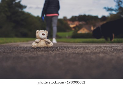 La muñeca de oso de Teddy perdida sentada en el sendero con perro borroso y mujeres caminando detrás con luz dramática, juguete de oso moreno solitario con cara triste mirando al parque público, Día Internacional de los Niños faltando