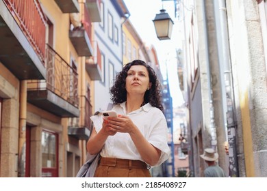 Lost Woman In Casual Clothes Walking On An Old European Street, Looking Around, Using A Map On Her Smartphone 