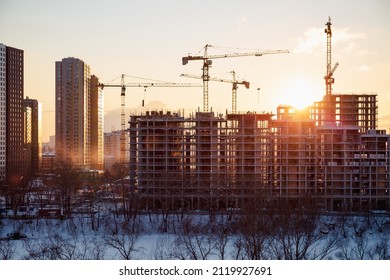 Lots of tower cranes build high-rise residential neighborhood on bank of frozen river in evening time. Industrial concept with sun flare. Yellow light and blue shadows.