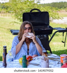 LOUISIANA / USA - JULY 15, 2017: A Young Woman Is Biting Into A Hamburger, Sitting Outdoors, In Front Of A BBQ Pit At A Table Full Of Condiments, On A Hot Summer Day.