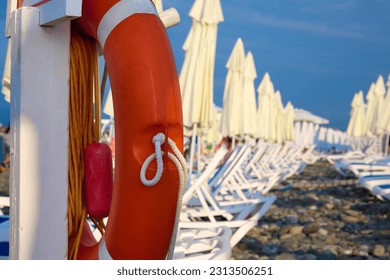 Lounge chairs with umbrellas lined up on clear rocky shore by sea against blue sky on beautiful beach. Lifebuoy and modern beach furniture located near the hotel poison with the sea for vacationers - Powered by Shutterstock