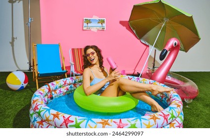 Lounging in colorful inflatable pool, slim young woman enjoys sun on neon-green float ring and drinks cold, refreshing soda. Concept of pop art, travel, party, recreation, lifestyle, fashion and style - Powered by Shutterstock