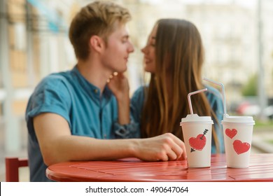 Love is in the air for two beloved ones who look on each other with passion in the blurred background. Two cups with coffee with red hearts and black arrows in the focus of the camera.