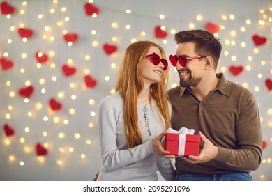 Love Background. Loving Couple In Festive Glasses Looks Into Each Other's Eyes Holding A Gift Box Sitting On A Background Of Hearts On The Wall. Love And Valentines Day Concept. Banner. Place For Text