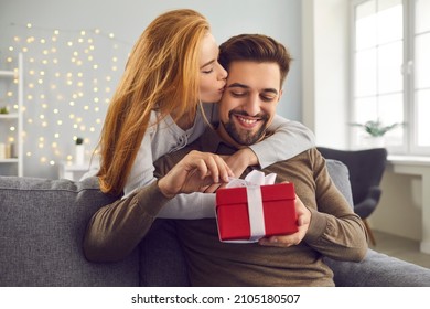 Love Background. Young Redhead Wife Hugs And Kisses Her Husband On The Cheek Giving Him A Gift For Valentine's Day. Smiling Bearded Man Unpacks His Gift While Sitting On Sofa At Home. Concept Of Love.