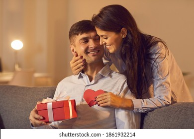 Love Background. Young Woman Stands Behind A Man Sitting On A Sofa And Gives Him A Gift And A Valentine For Valentine's Day. Family Celebrates At Home In A Cozy Room. Concept Of Love.