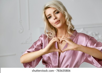 Love. Beautiful Female Model Showing Heart With Hands. Portrait Of Smiling Woman In Silk Pink Fashion Robe With Gorgeous Hair Style And Natural Makeup Making Heart Sign With Hands. High Resolution