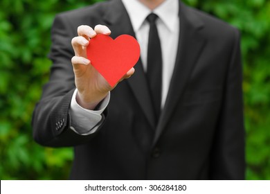 Love And Business Theme: A Man In A Black Suit Holding A Card In The Shape Of A Red Heart On The Background Of Green Grass