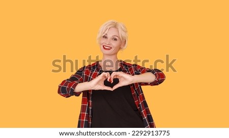 Love care. Affected woman. Feelings expression. Happy middle-aged lady holding hands forming heart shape gesture on yellow.