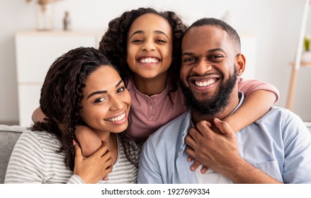 Love And Care. Portrait Of Cheerful African American Family Of Three People Hugging Sitting On The Sofa At Home, Posing For Photo And Looking At Camera. Smiling Young Girl Embracing Her Parents