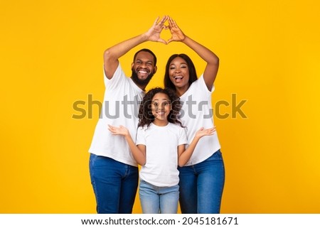 Love, Care And Support. Smiling black parents showing heart shape with fingers, connecting their hands together, making symbolic roof under their daughter head isolated on yellow orange studio wall