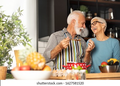 Love Couple Grandparents Cooking Together While Man Feeding His Wife At The Kitchen. Long Lasting Relationship Concept, Valentine’s Day