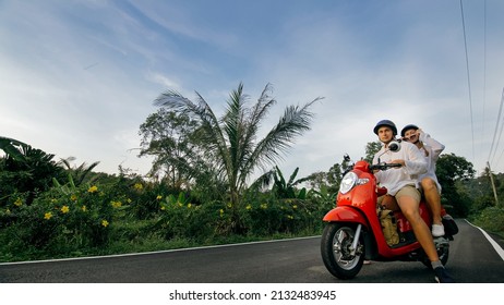 Love Couple On Red Motorbike In White Clothes On Forest Road Trail Trip. Two Caucasian Tourist Woman And Man Kiss Hugs Sit On Scooter. Motorcycle Rent Safety Helmet Sunglasses. Asia Thailand Tourism.