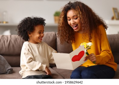 With love  Cute excited little boy son giving his mom handmade greeting postcard and red heart  happy mother embracing and with child while sitting sofa at home  Mothers day celebration concept