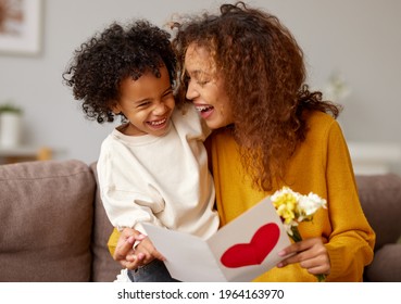 With love. Cute excited little boy son giving his mom handmade greeting postcard and flower bouquet, happy mother embracing with with child while sitting on sofa at home. Mothers day  concept