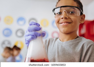 Love experimenting. Cheerful teenage boy in safety glasses smiling at the camera while holding a chemical flask and watching a chemical reaction in it
