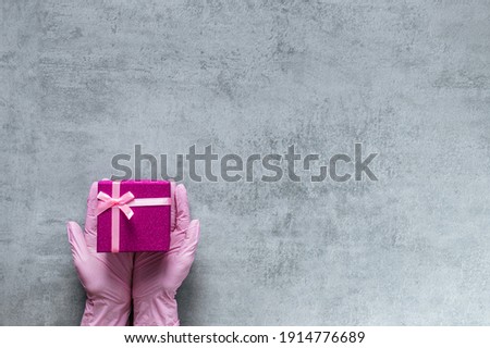Love gift. Covid-19 protection. Hands in medicine glove holding pink present box on grey copy space. Advertising background. Valentine day greeting. A gift for a woman