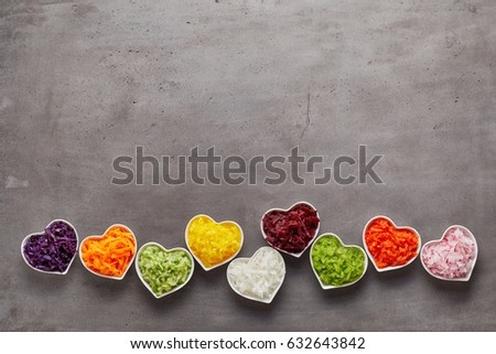 Love for healthy food concept with little heart-shaped white bowls of grated vegetables of different colors in a row on grey surface background with copy space