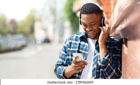 I Love It. Excited african guy touching headphones and holding cell phone, listening to song, leaning on the wall