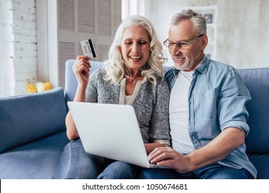 Love Lives Forever! Senior Couple At Home. Handsome Old Man And Attractive Old Woman Are Spending Time Together. Sitting On A Sofa With Laptop And Credit Card In Hands. Online Shopping