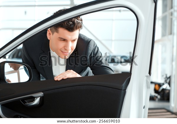 Love luxury cars. Smiling\
handsome man in formalwear looking inside the new car in car\
dealership