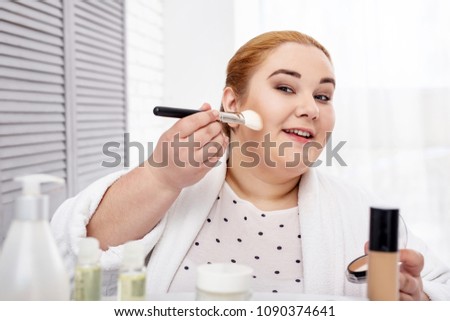 I love makeup. Smiling stout woman wearing a bathrobe and applying make-up