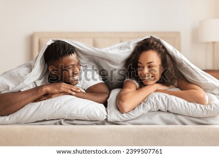 Love And Marriage. Young African American Couple Lying Under Blanket In Bed, Enjoying Intimate Moment, Smiling To Each Other Flirting In Modern Bedroom. Morning Together Concept