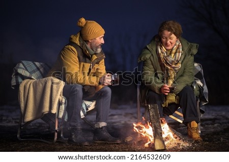 In love mature couple romantic dinner at campfire drinking hot natural tea sitting on the plaid blankets in winter journey. Couple sitting at campfire in snow. Family vacation and journey concept. 