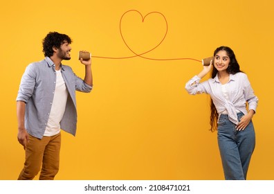 Love Message. Loving Indian Man Telling Romantic Words To Girlfriend Through Tin Can Phone With Heart Shaped String, Standing Over Yellow Background, Creative Collage