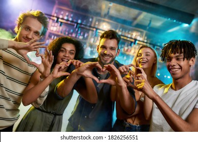 Love my friends, young men and women showing heart signs while posing together for camera, multiracial group of friends hanging out at party in the bar. Focus on hands.