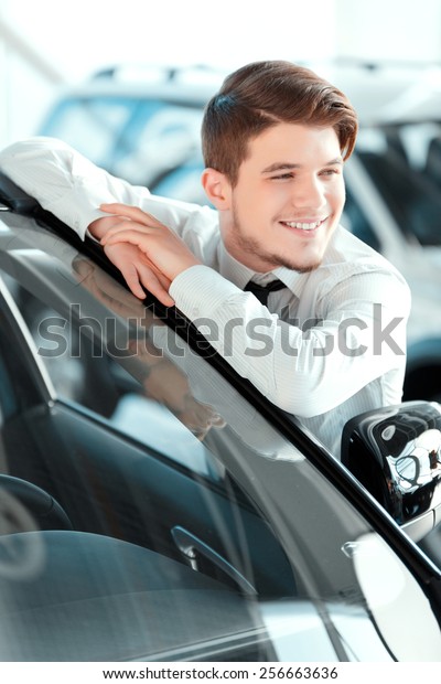 I
love my job. Closeup of handsome young car salesman in formal wear
standing at the dealership and leaning at the car 

