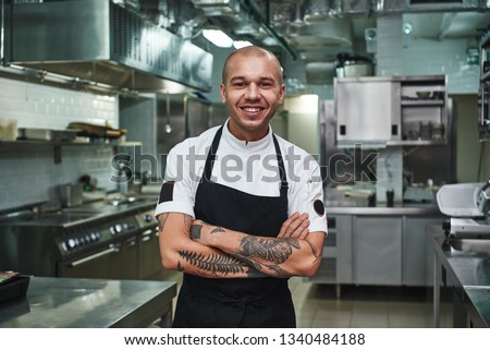 I love my work Cheerful young chef in apron keeping tattooed arms crossed and smiling while standing in a restaurant kitchen