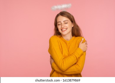 I love myself. Portrait of gorgeous angelic ginger girl with halo over head embracing herself lovingly and smiling from pleasure, self-esteem concept. indoor studio shot isolated on pink background