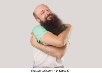 I Love Myself. Portrait Of Happy Middle Aged Bald Man With Long Beard In Light Green T-shirt Standing Hugging Himself Smiling And Enjoying. Indoor Studio Shot, Isolated On Grey Background.