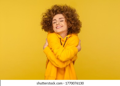 I love myself! Portrait of self-satisfied egoistic curly-haired woman in urban style hoodie embracing herself and smiling with pleasure, feeling self-pride. studio shot isolated on yellow background
