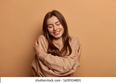 I love myself. Pretty smiling woman embraces herself gently, expresses self love, keeps eyes closed with pleasure, feels comfortable and fullfilled, being egoistic person, isolated on brown background