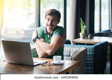 I Love Myself! Young Happy Satisfied Businessman In Green T-shirt Sitting, Hugging Himself And Looking At Camera With Toothy Smile. Business And Freelancing Concept. Indoor Shot Near Window At Daytime