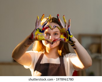 Love Painting. Female Artist. Creative Lifestyle. Art Studio. Inspiration Muse. Happy Artistic Woman In Apron Showing Heart Sign Hands With Colorful Paints Light Room Interior.