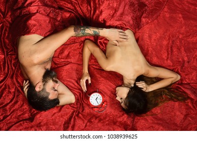 Love and perfect morning concept. Couple in love on red sheets. Man and woman with half covered bodies lie near red alarm clock. Guy with beard and tattoos touches pretty ladys back in bed, top view
