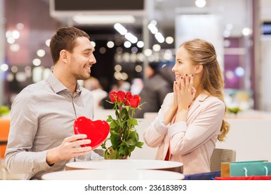 Love, Romance, Valentines Day, Couple And People Concept - Happy Young Man With Red Flowers Giving Present To Smiling Woman At Cafe In Mall