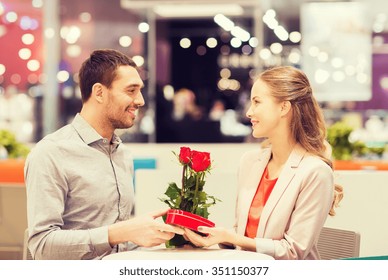 Love, Romance, Valentines Day, Couple And People Concept - Happy Young Man With Red Flowers Giving Present To Smiling Woman At Cafe In Mall