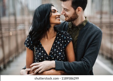 Love story in New York. Gorgeous couple of American man with beard and tender Eastern woman hug each other before the cityscape of Brooklyn bridge somewhere in New York