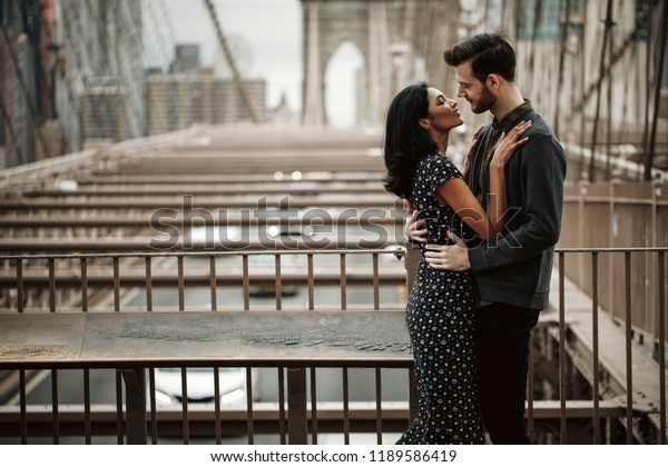ニューヨークのラブストーリー ニューヨークのどこかのブルックリン橋の都市の風景の前で あごひげを生やし 優しい東洋の女性を持つ華麗な2人のアメリカ人男性が抱き合う の写真素材 今すぐ編集