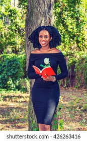Love Story. Wearing long sleeve, slim, off shoulder dress, holding red book with white rose, African American woman with braid hairstyle standing by tree on park in New York, reading, smiling.