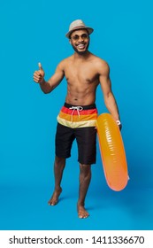 Love summer. Friendly african guy showing thumb up, going with inflatable ring on blue studio background
