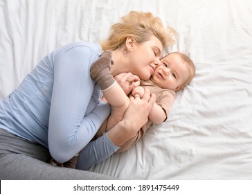 Love and tenderness, motherhood concept. Loving blonde mother caressing her cute baby boy, lying together on bed, top view. Young caucasian woman bonding with little kid at home, copy space