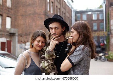 Love triangle between man and two women. Outdoor shot of bearded hipster in trendy hat looking at camera with thoughtful expression, choosing between beautiful females who standing next to him