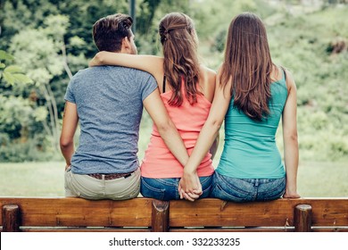 Love triangle, a girl is hugging a guy and he is holding hands with another girl, they are sitting together on a bench